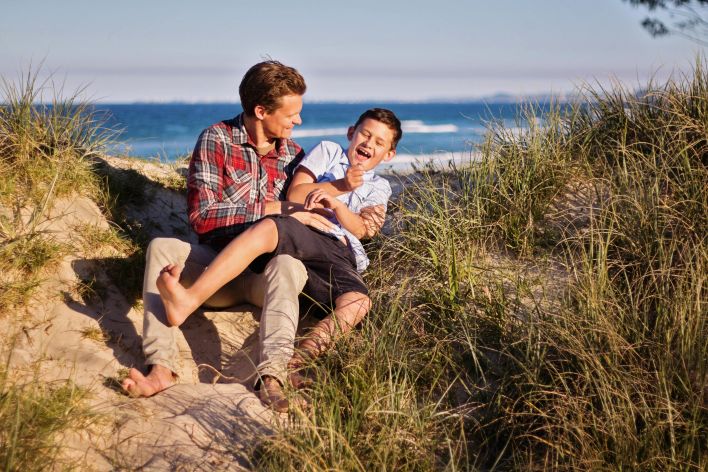 A joyful father and son share a laugh on a sunny beach, surrounded by sand dunes.