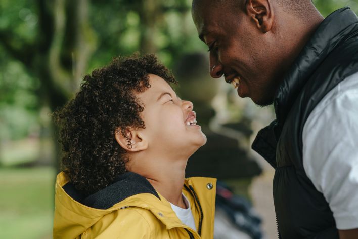 Side view of African American young father smiling while looking on joyful little son with closed eyes on blurred background